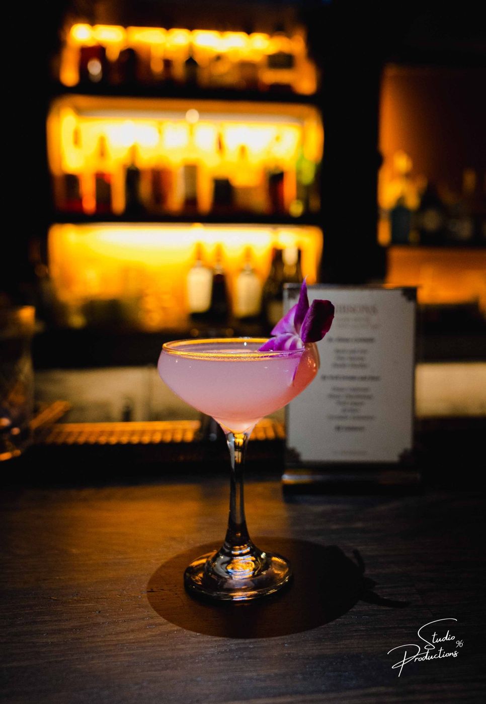 A pink cocktail garnished with an orchid flower on the bar