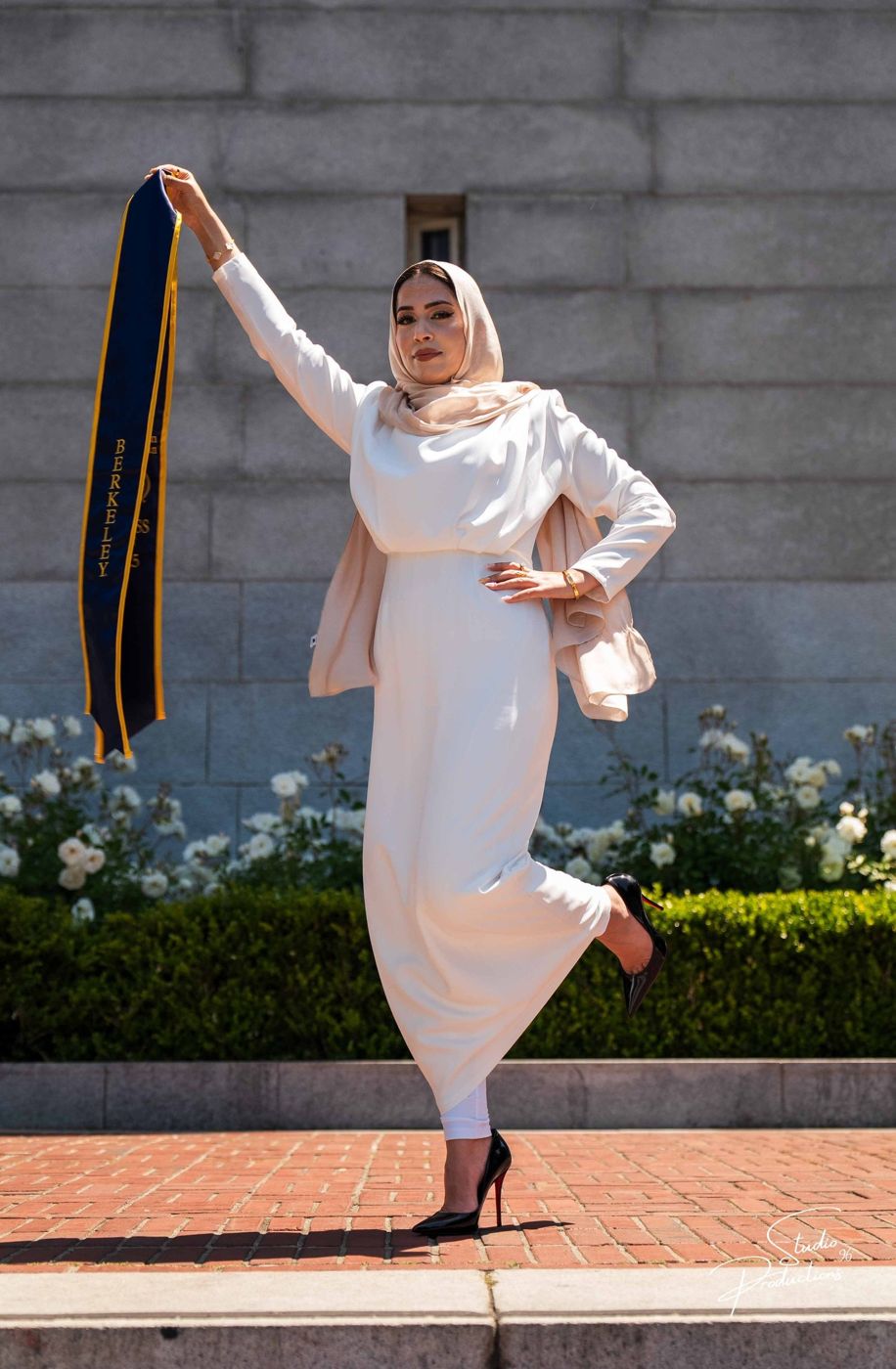 Graduate in a cream dress striking a playful pose, holding up her graduation sash
