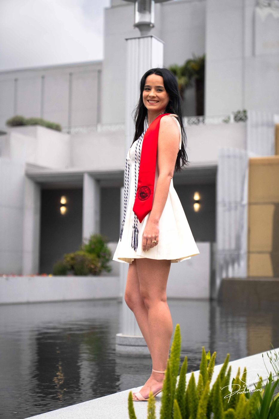 Graduate in a white dress and sash standing by a reflecting pool