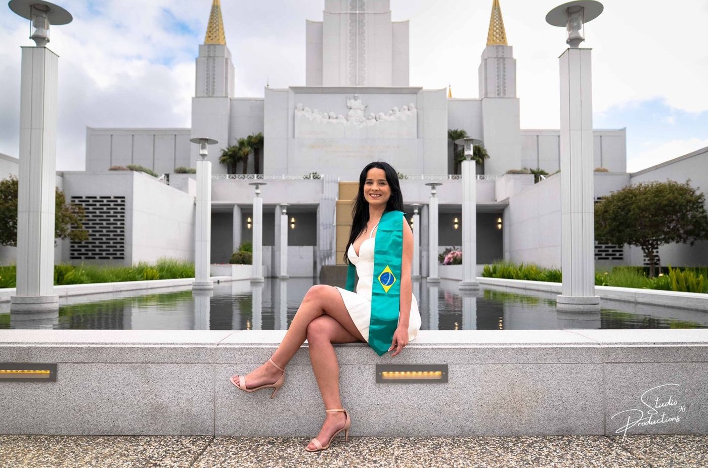 Graduate in a white dress and sash seated by the reflecting pool