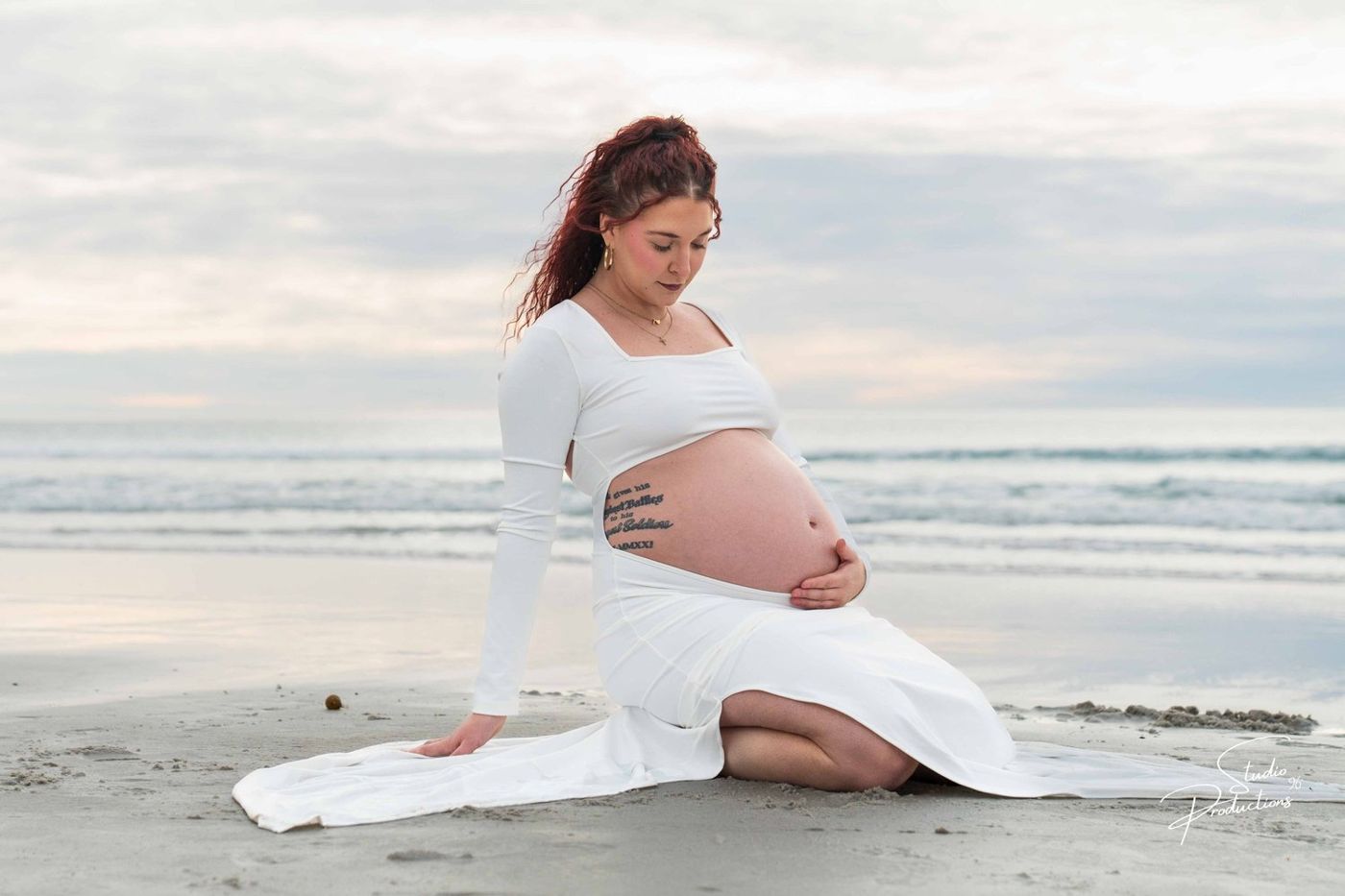 Expecting mother sitting on the sand at the beach in a white gown, looking down at her belly