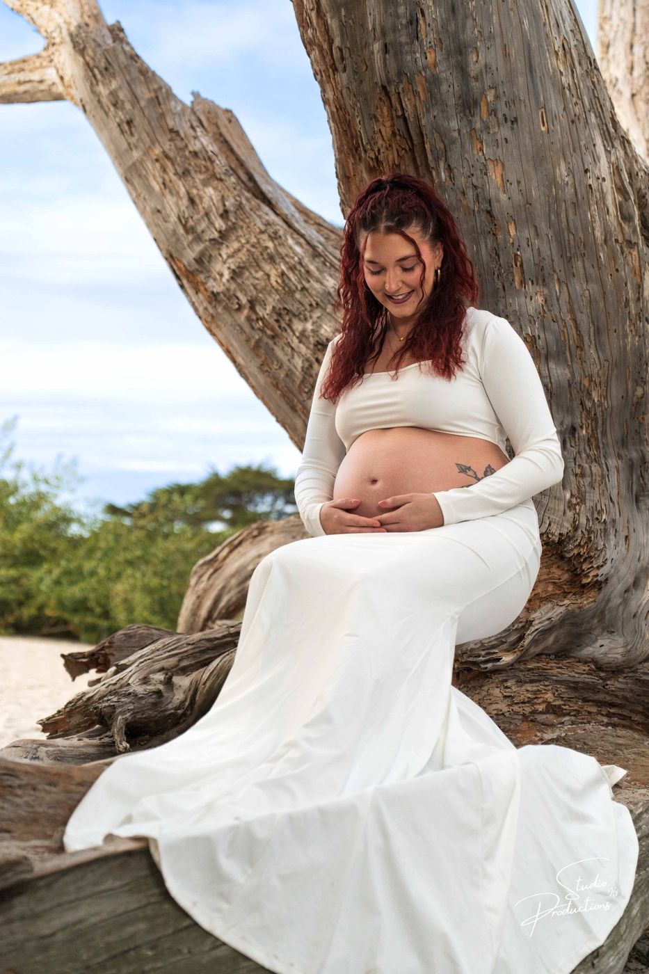 Expecting mother sitting against a large driftwood trunk on the beach, cradling her belly