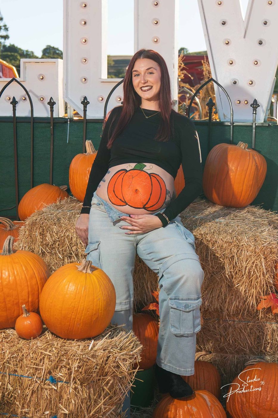 Expecting mother sitting on a hay bale with a pumpkin painted on her belly, beneath a lit 'LOVE' sign at the pumpkin patch