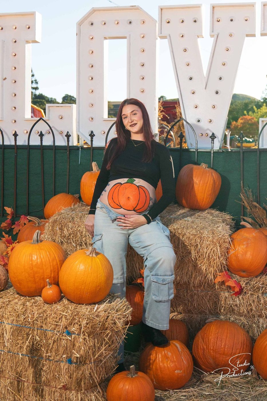 Expecting mother framing her painted-pumpkin belly with both hands at the 'LOVE' sign pumpkin patch