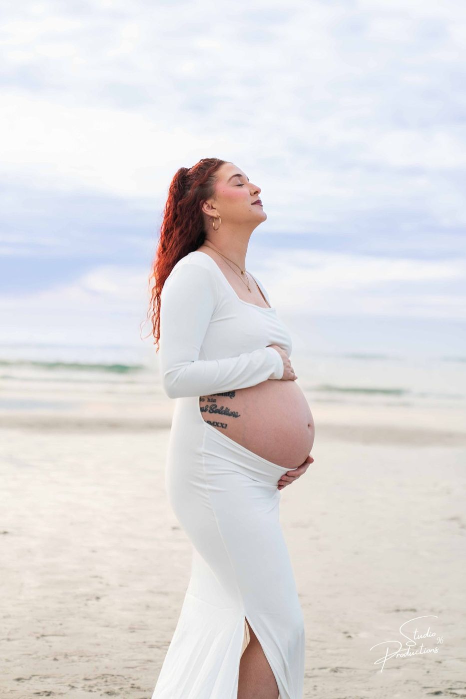 Expecting mother standing on the beach, looking up at the sky, cradling her belly