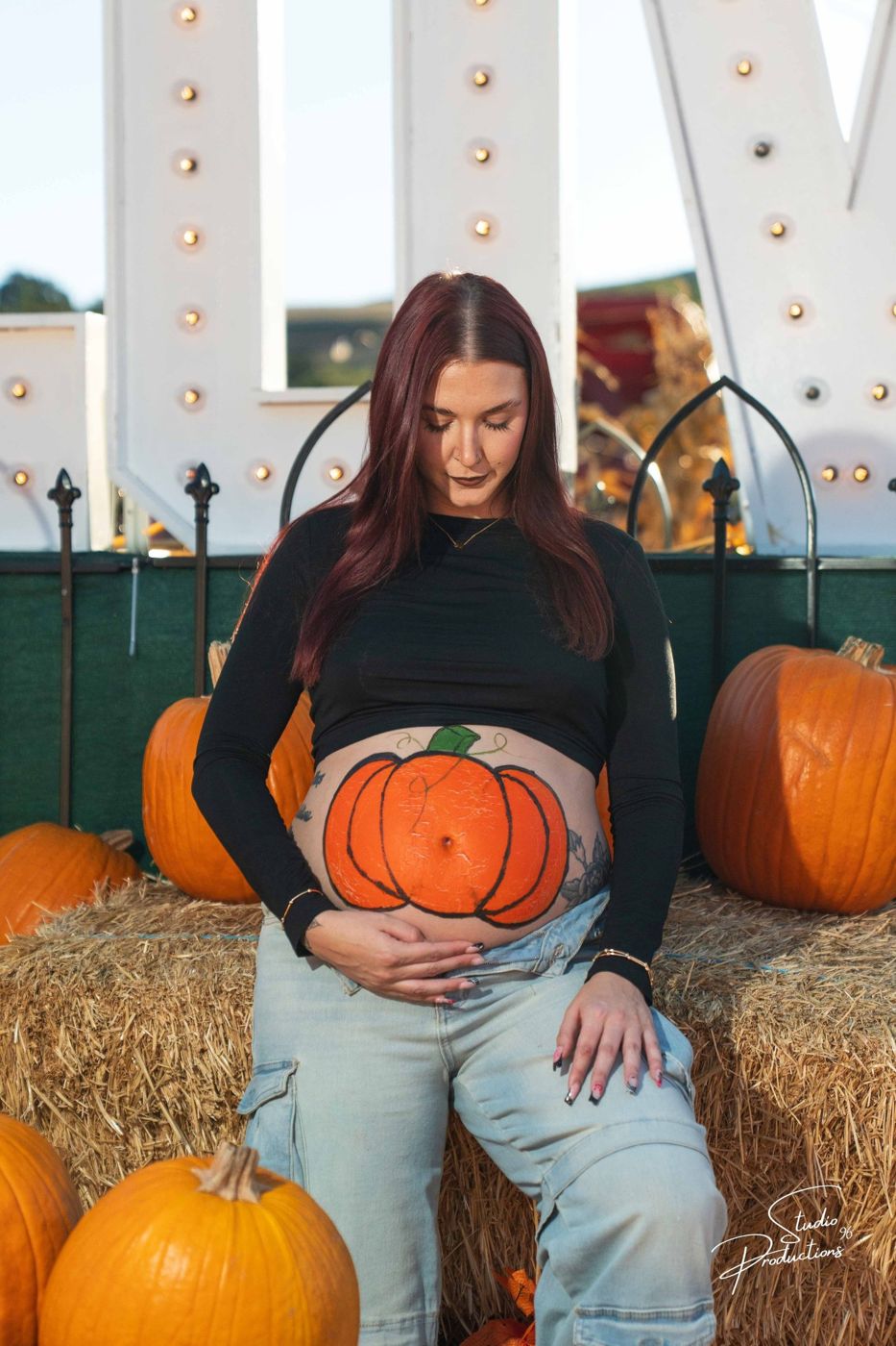 Expecting mother sitting on a hay bale, looking down at the pumpkin painted on her belly