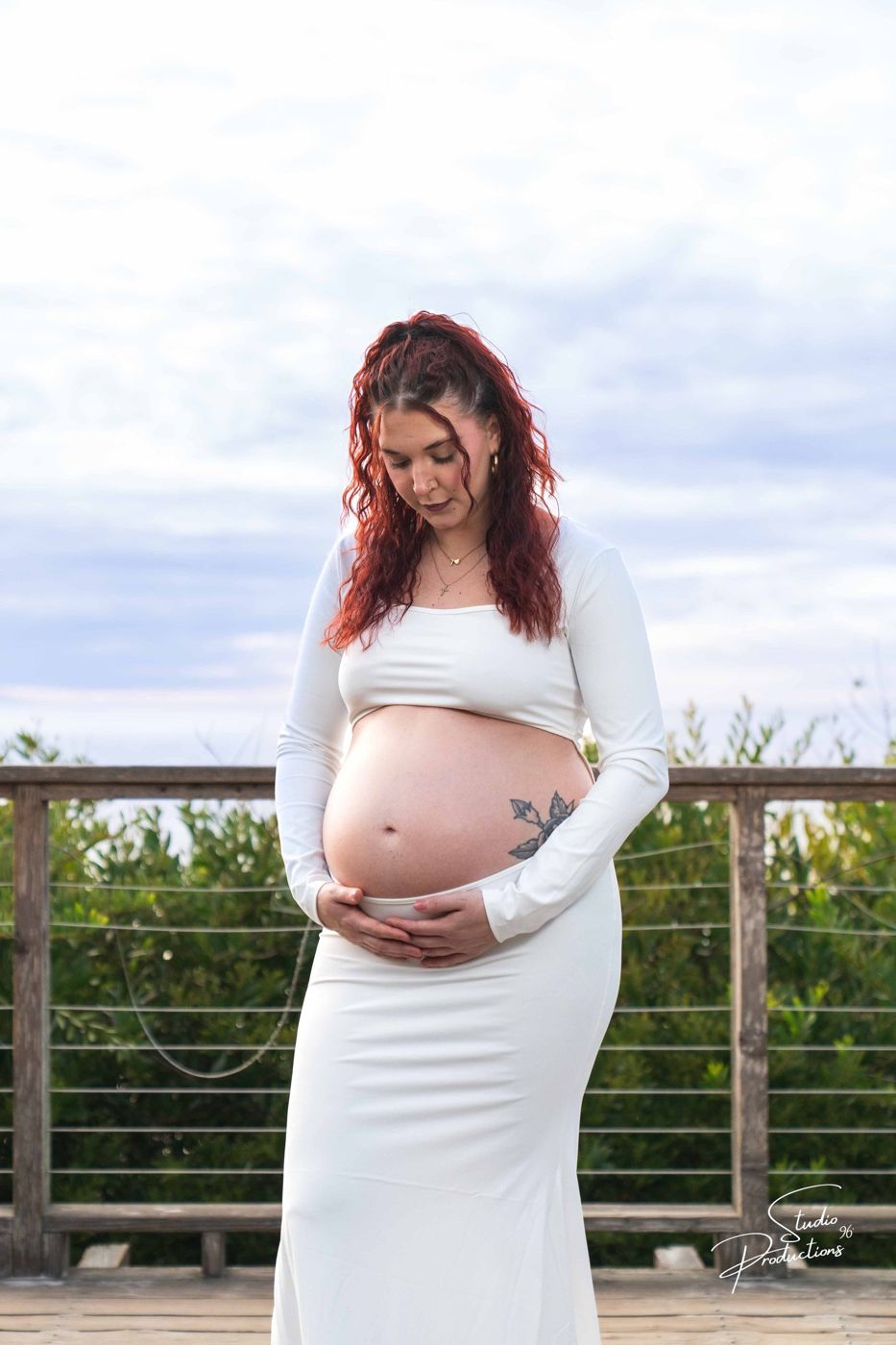Expecting mother standing at a waterfront railing in a white dress, cradling her belly