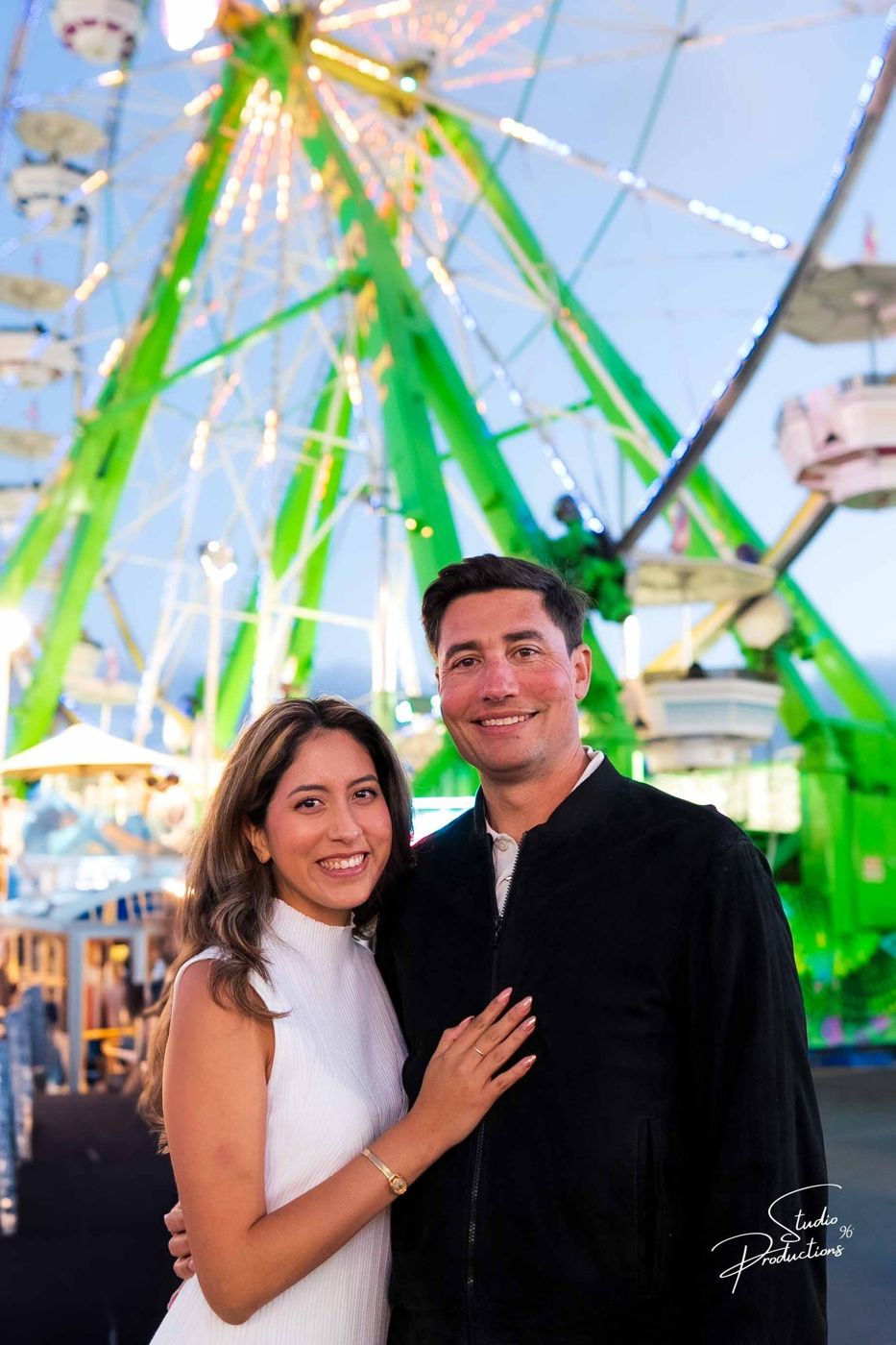 Couple standing together on a rock formation, a lit ferris wheel behind them at dusk