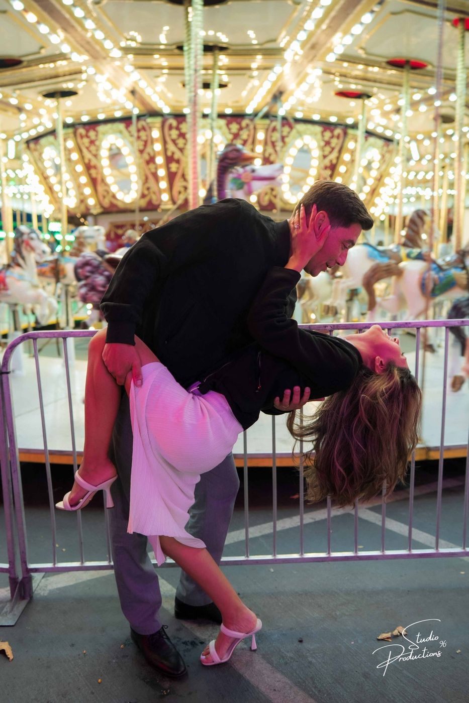 A dramatic dip-kiss pose at the carousel, colorful lights behind them