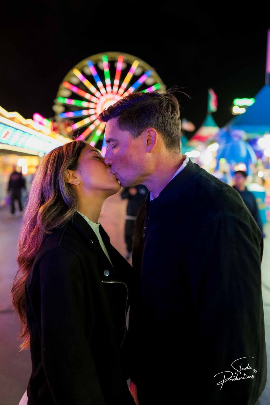 Close-up of the couple kissing at night, a ferris wheel glowing behind them