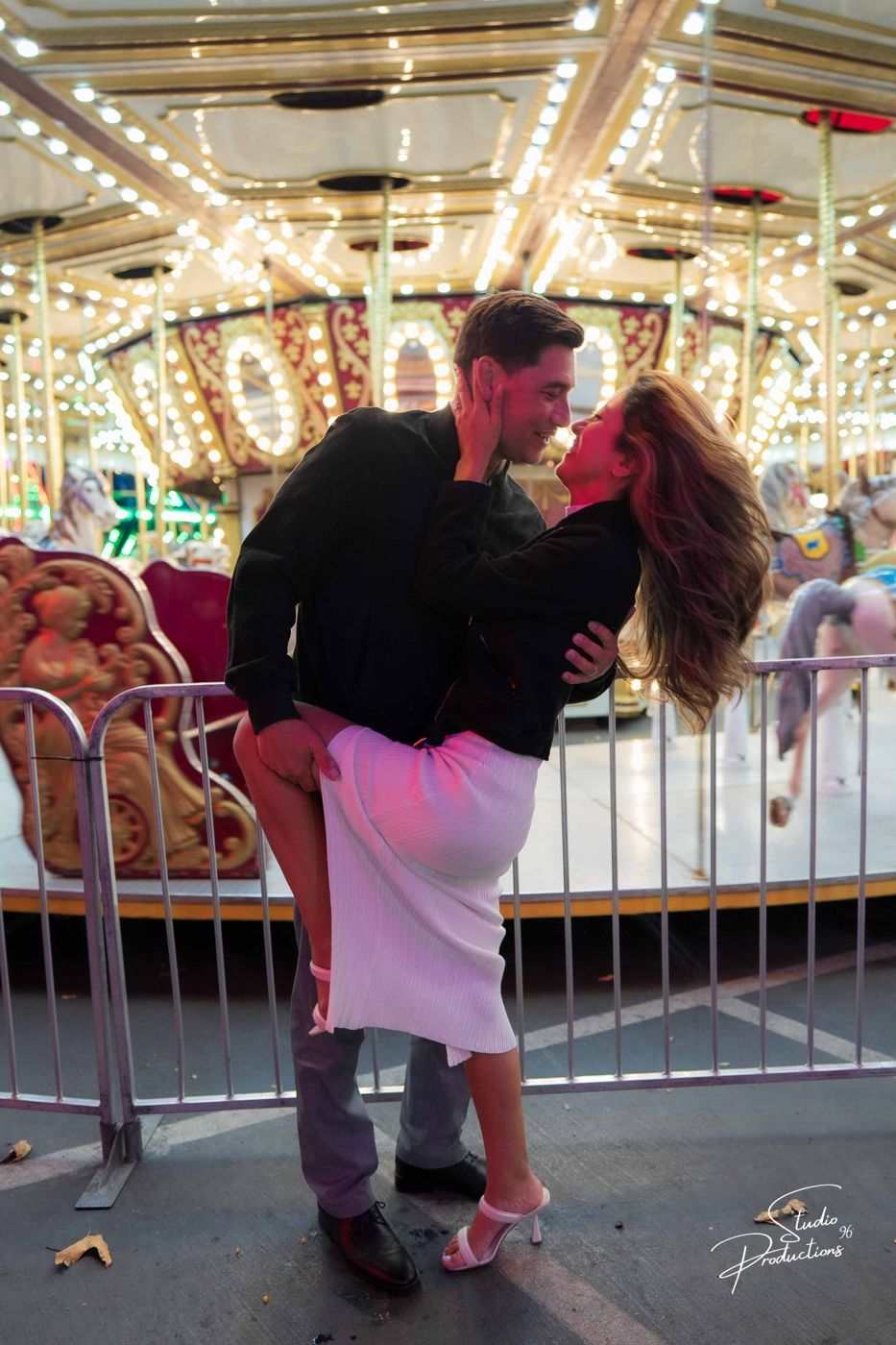 A dramatic dip-kiss at the carousel, colorful ride lights behind them