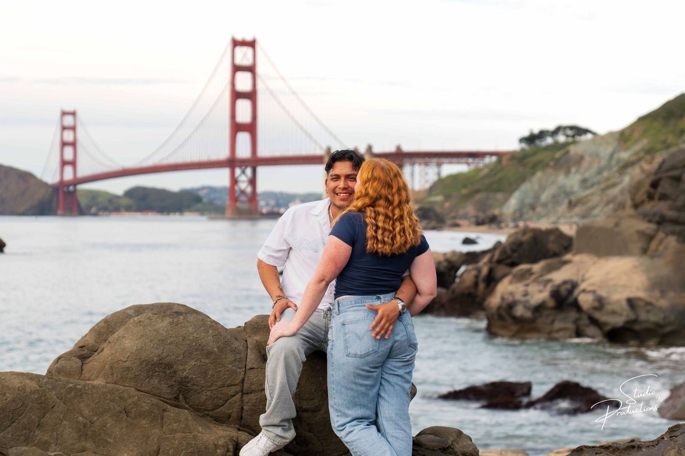 Couple standing on rocks along the coastline with the bridge behind them